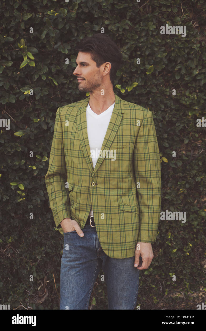 A good looking Caucasian man standing in front of a green bush, wearing a green plaid 70s jacket and jeans, standing and looking away. Stock Photo
