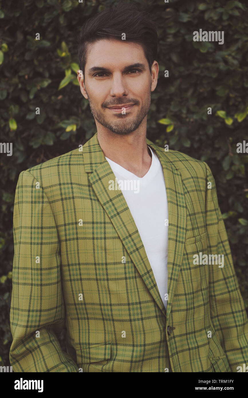 A portrait of handsome male model wearing a vintage 70s jacket, looking happy and friendly. A close-up portrait in front of a green bush background. Stock Photo