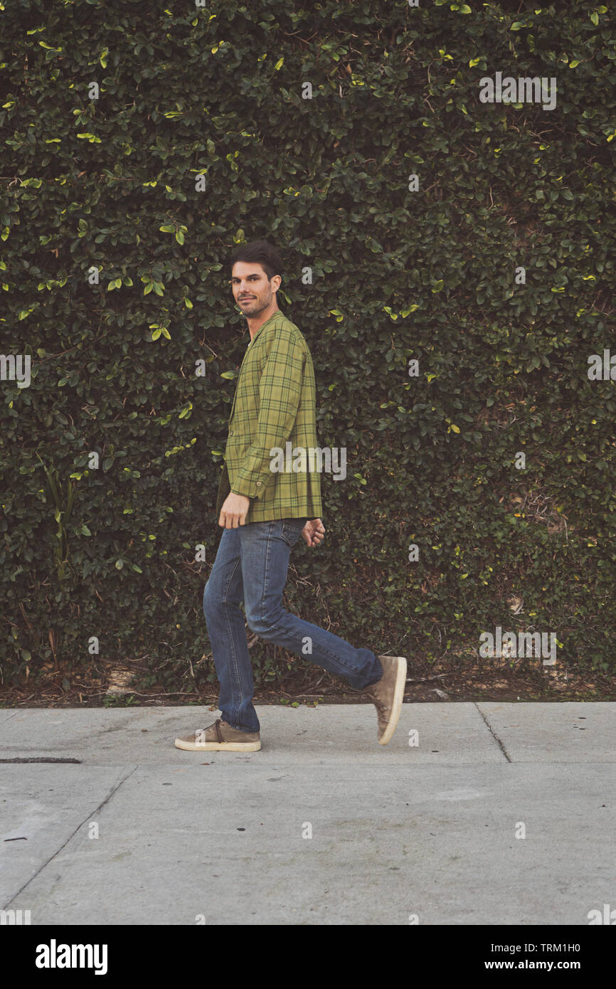 A male model, man walking on a pavement in front of a large green bush wall. Stock Photo