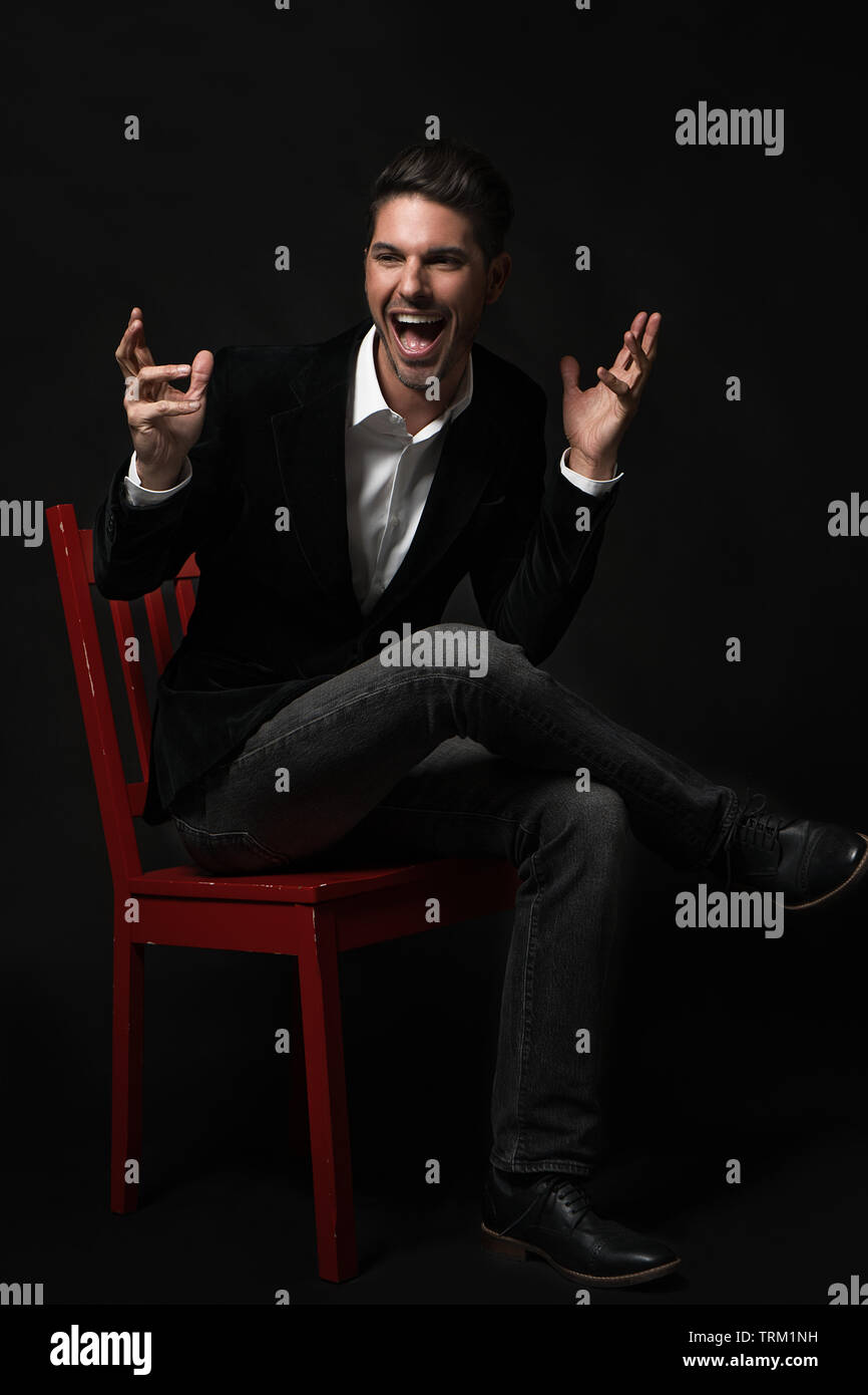 A good looking white male model modeling in a formal jacket, sitting on a red chair crossed legs. He throws his hands up in excitement and screaming! Stock Photo
