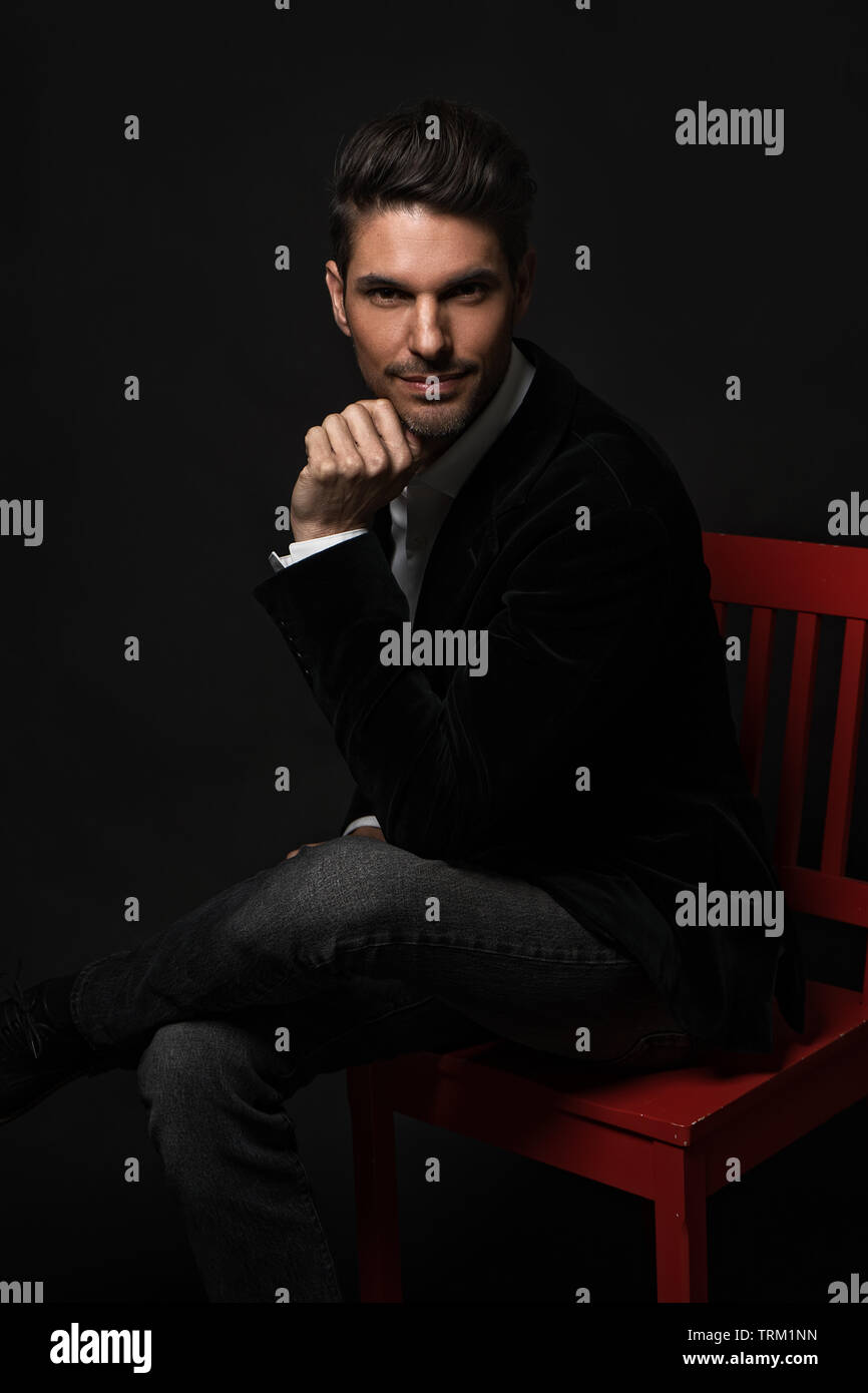 A good looking white male model modeling in a formal attire, sitting on a red chair crossed leg, hand to chin, looking at the camera with confidence. Stock Photo