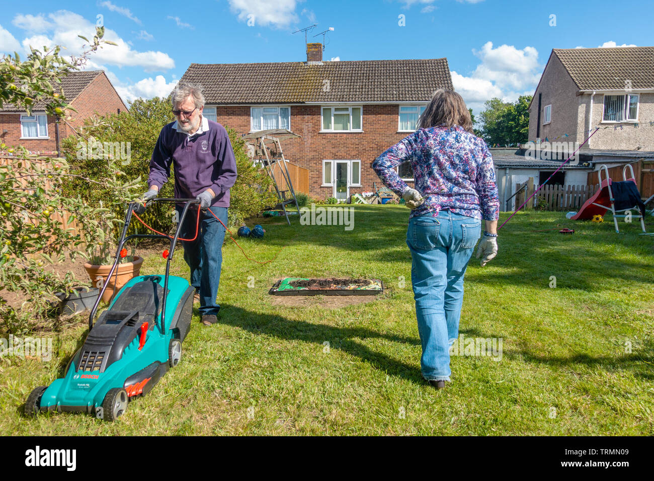A retired couple doing gardening in a residential back garden on a sunny day with blue sky. The man mows the lawn with an electric lawnmower. Stock Photo