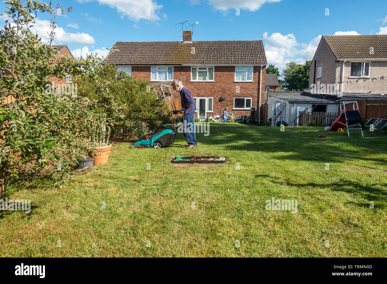 A man mows a lawn in a residential back garden with an electric lawnmower in a sunny day with blue sky in early summer. Stock Photo