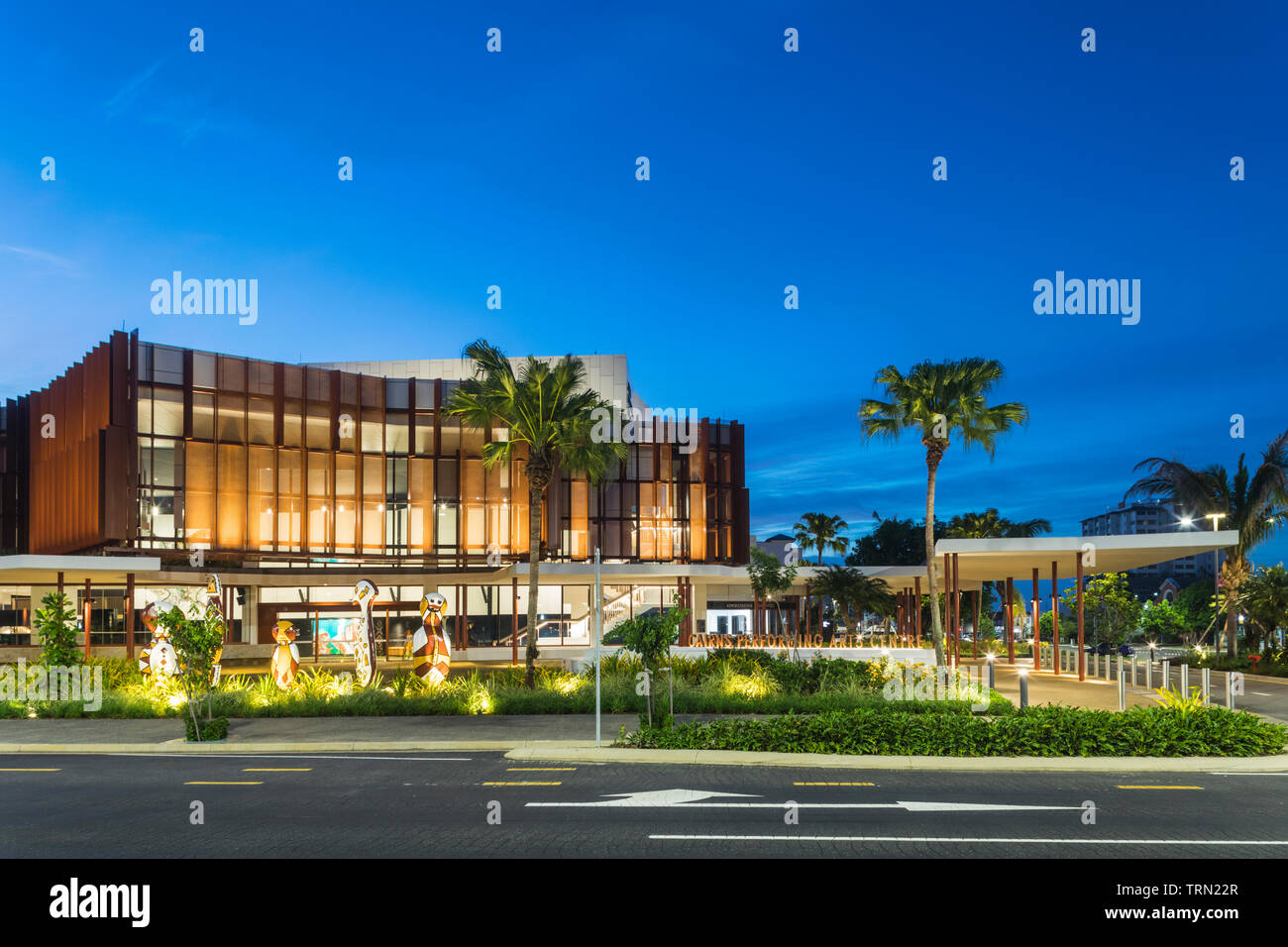The facade of the Cairns Performing Arts Centre illuminated at twilight.  A set of 'Bagu' indigenous sculptures sit at the front of the centre.  Cairn Stock Photo