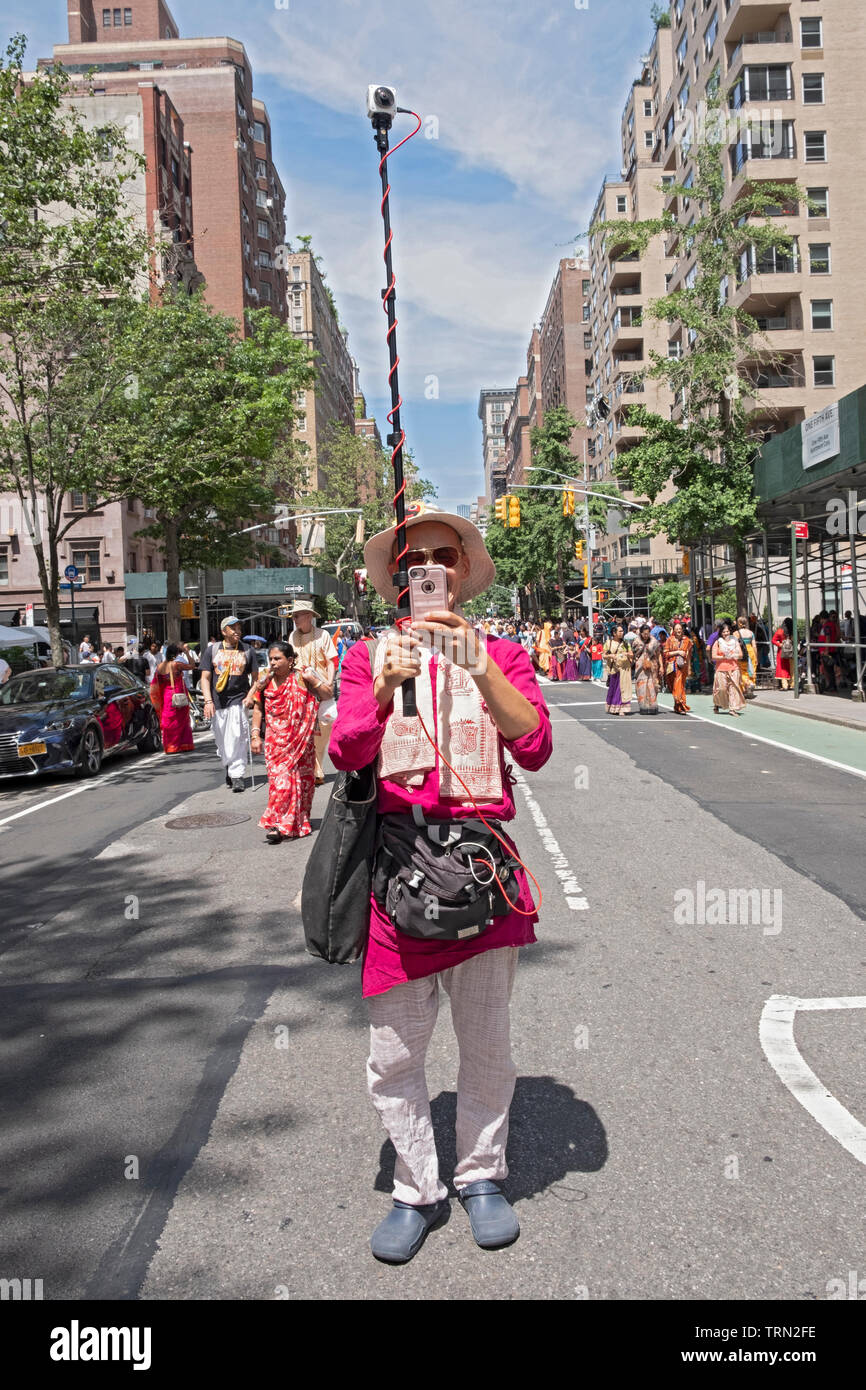 A man at the front of the Hare Krishna Ratha Yatra parade in NYC both takes a selfie & photographs the parade with a go pro camera pointed backwards. Stock Photo