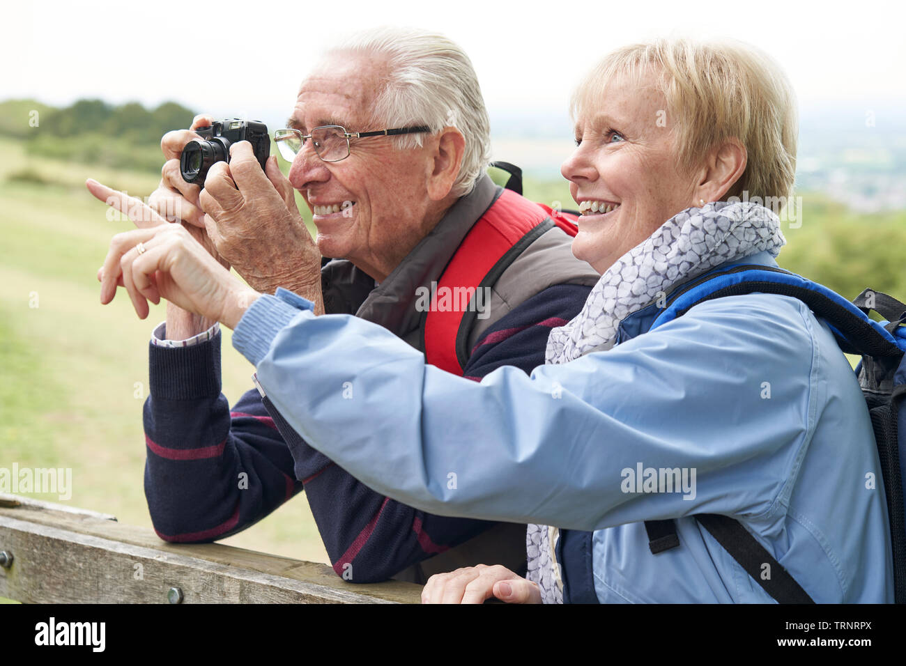 Senior Couple Hiking In Countryside Standing By Gate  And Taking Photo With Camera Stock Photo