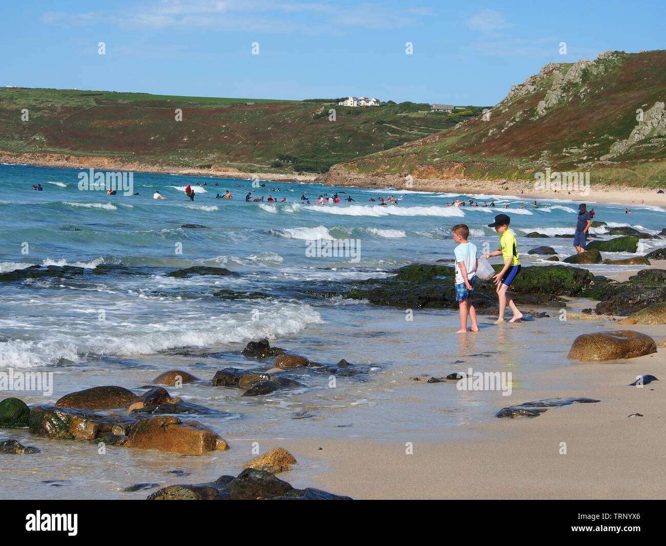 People enjoying a sunny summers day on the beach in Sennen Cove, Cornwall, England, UK showing the sandy beach and blue sea under a blue sky. Stock Photo
