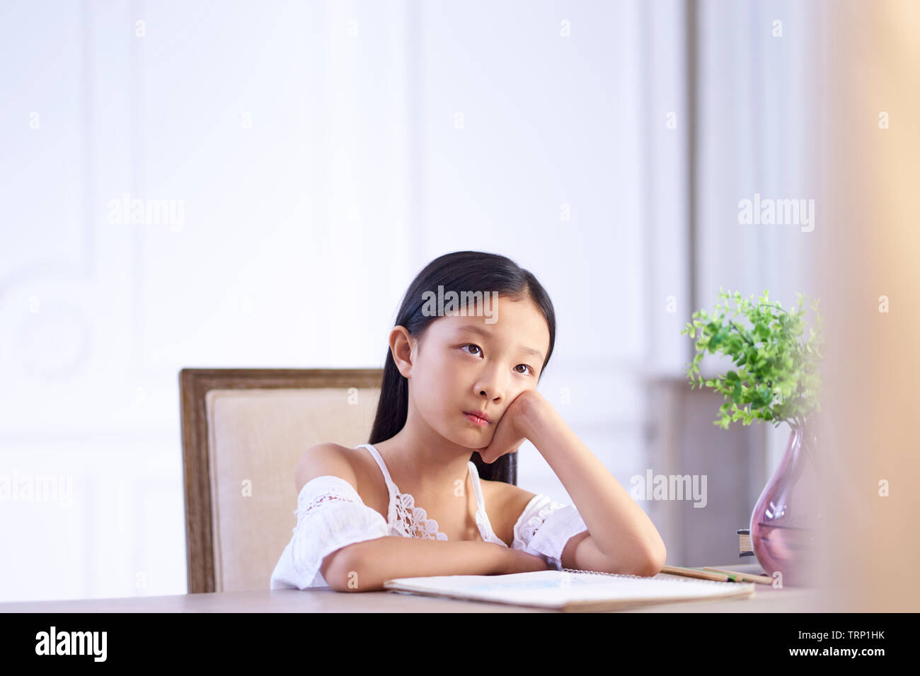 9 year-old little asian girl sitting at desk thinking with hands on cheek. Stock Photo
