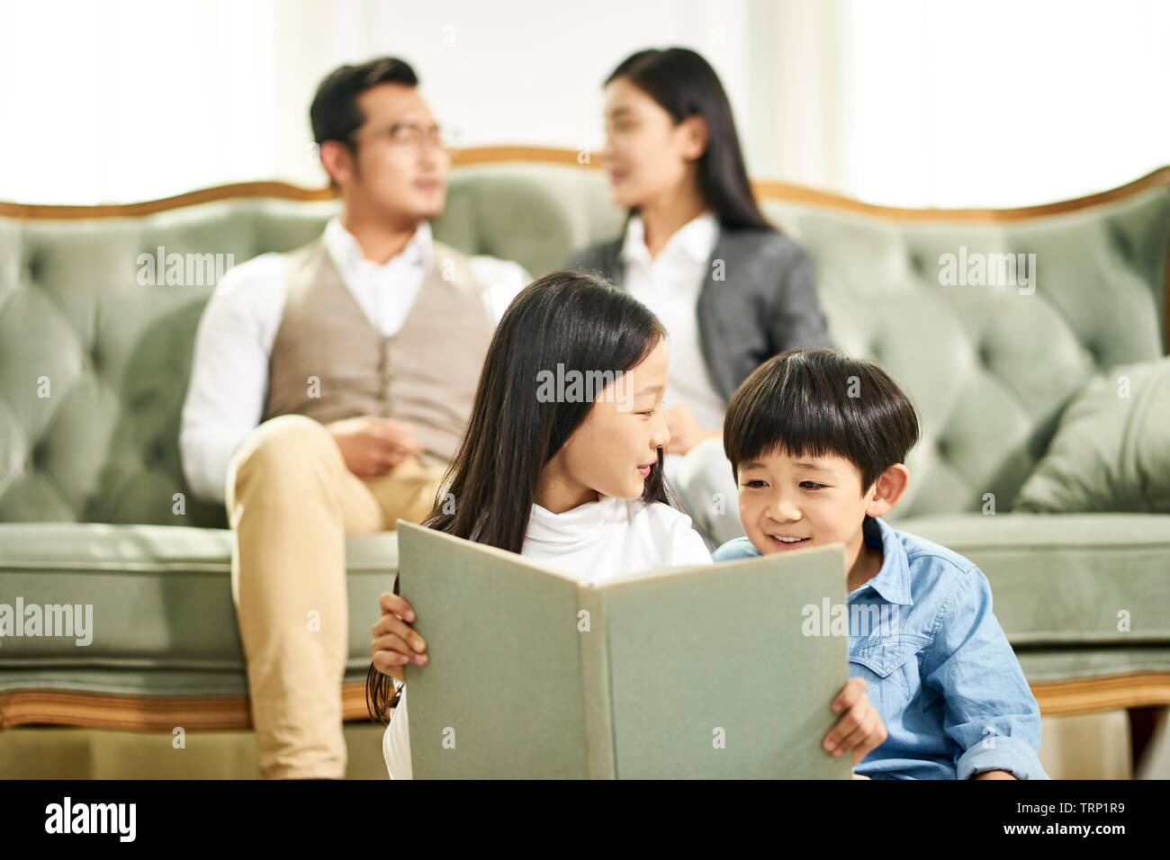 asian brother and sister sitting on carpet in family living room reading book together while parents talking in background. Stock Photo