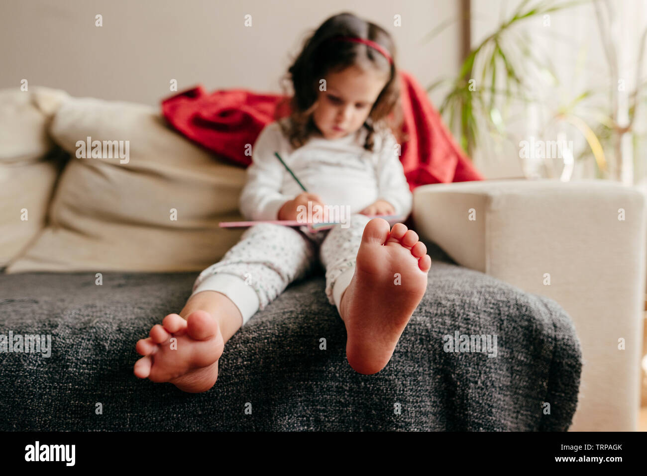 Unfocused barefoot little girl sitting on the couch and drawing in the notebook Stock Photo
