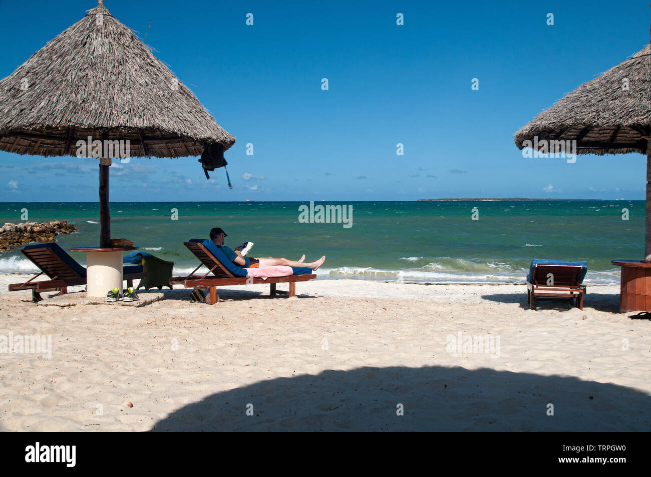 A relaxing tourist is reading a book by the side of sea beach in Dares Salaam,Africa, stretching his legs on an easy chair with his shoes off. Stock Photo