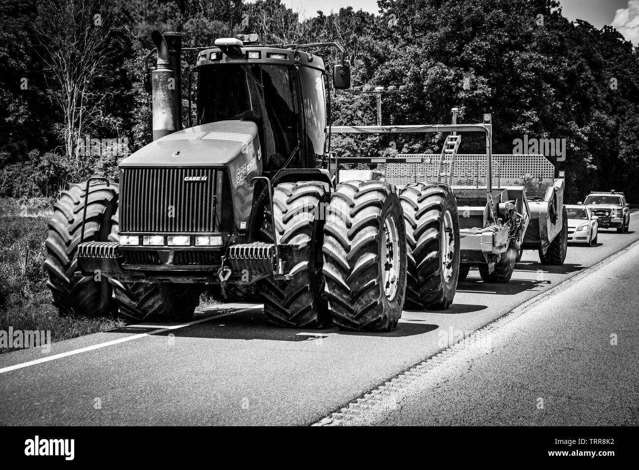 A huge, modern, farm tractor, a definite wideload, is holding up traffic as it travels down a rural highway in Northwestern Mississippi, in black and Stock Photo