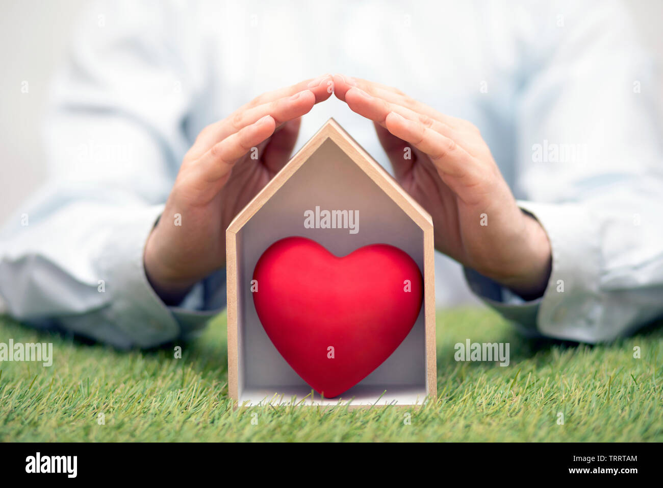 Wooden house with red heart on green grass protected by hands Stock Photo