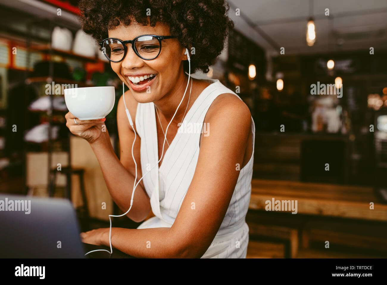 Young african woman drinking coffee and using laptop at a cafe. Smiling woman sitting at cafe drinking coffee and looking at laptop on table. Stock Photo