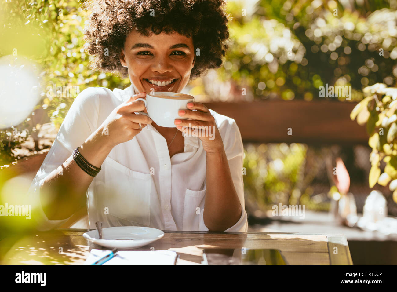 Cheerful young african woman with a cup of coffee sitting at cafe. Female having coffee at coffeeshop. Stock Photo
