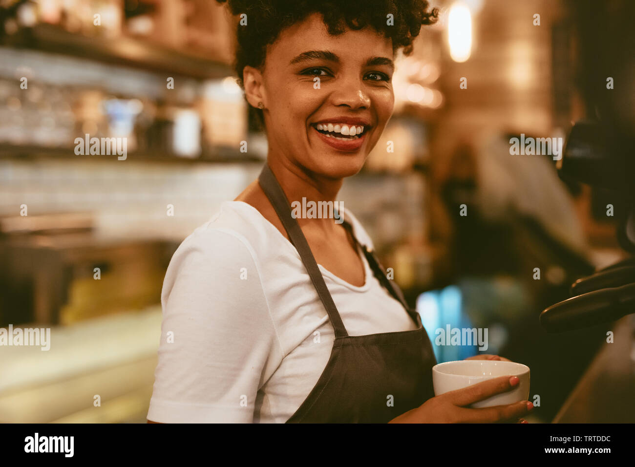 Female barista holding a cup standing by a coffee maker. Female cafe worker preparing coffee in machine. Stock Photo