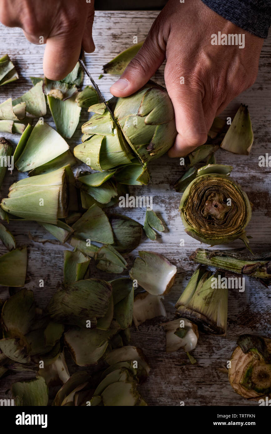 closeup of a caucasian man cutting with a kitchen knife some raw fresh artichokes, on a white rustic wooden table Stock Photo