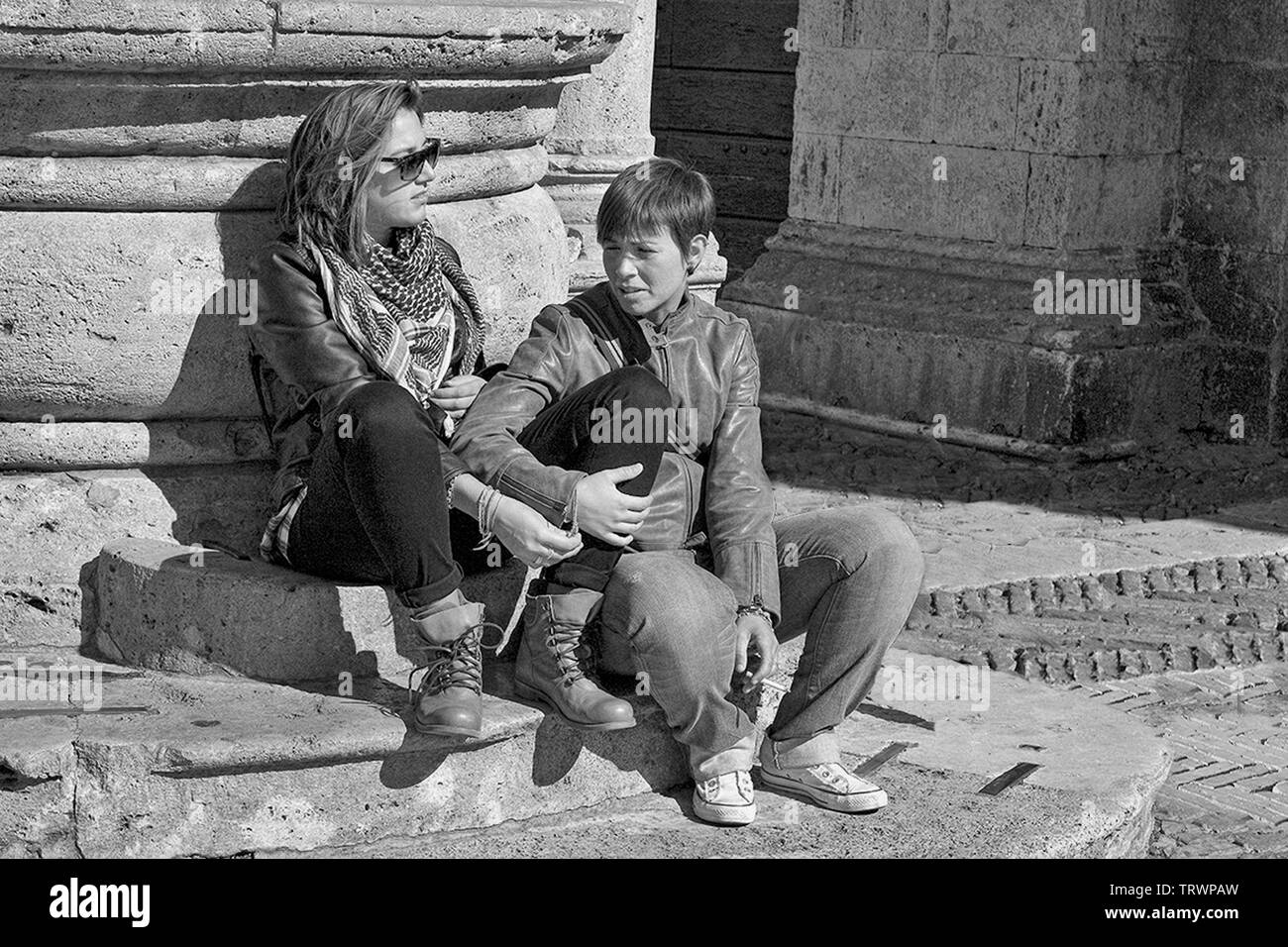 Two girls sit at the base of the fountain, known as Pozzo dei Grifi e dei Leoni in the Piazza Grande, Montepulciano, Tuscany, Italy.  Black and white Stock Photo