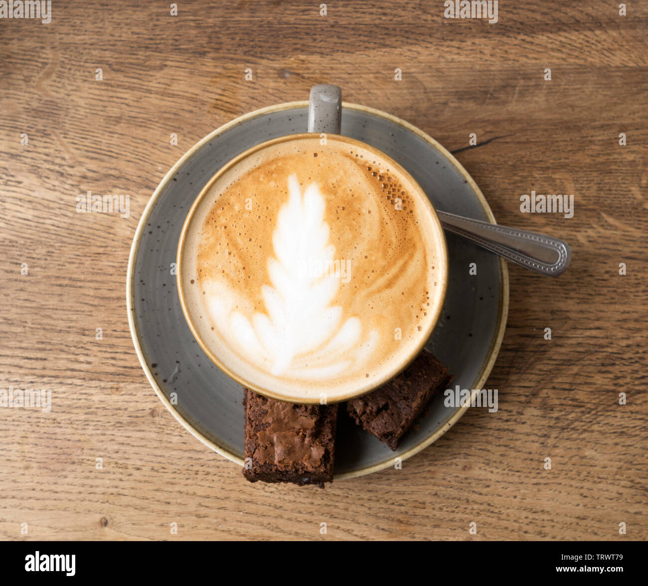 A cup of flat white coffee with chocolate brownies and a spoon on a saucer against a wood effect background, England, UK Stock Photo