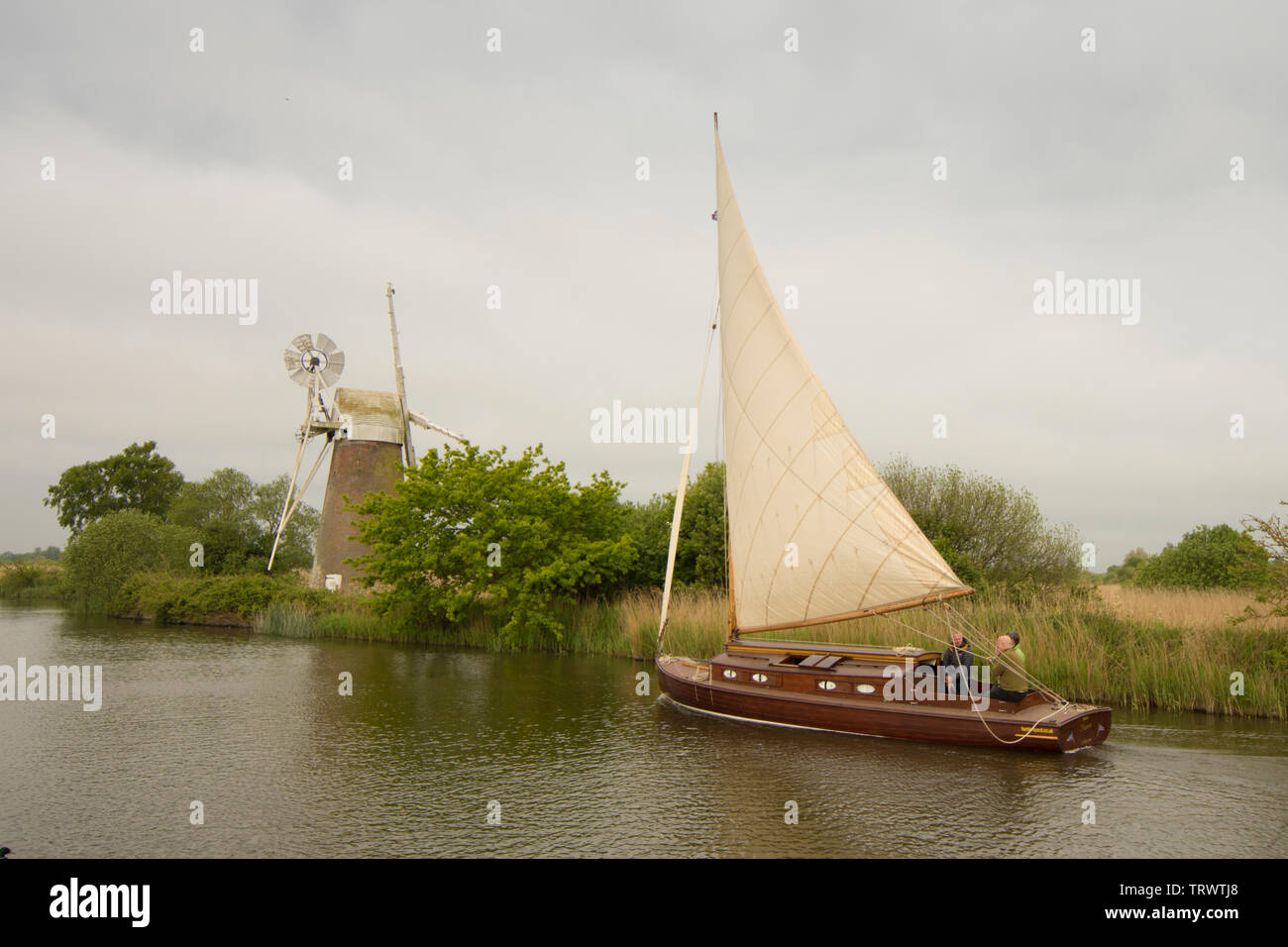 Three men in sailing boat on River Ant sailing in front of Turf Fen Drainage Mill, How Hill, Norfolk Broads, UK, May Stock Photo