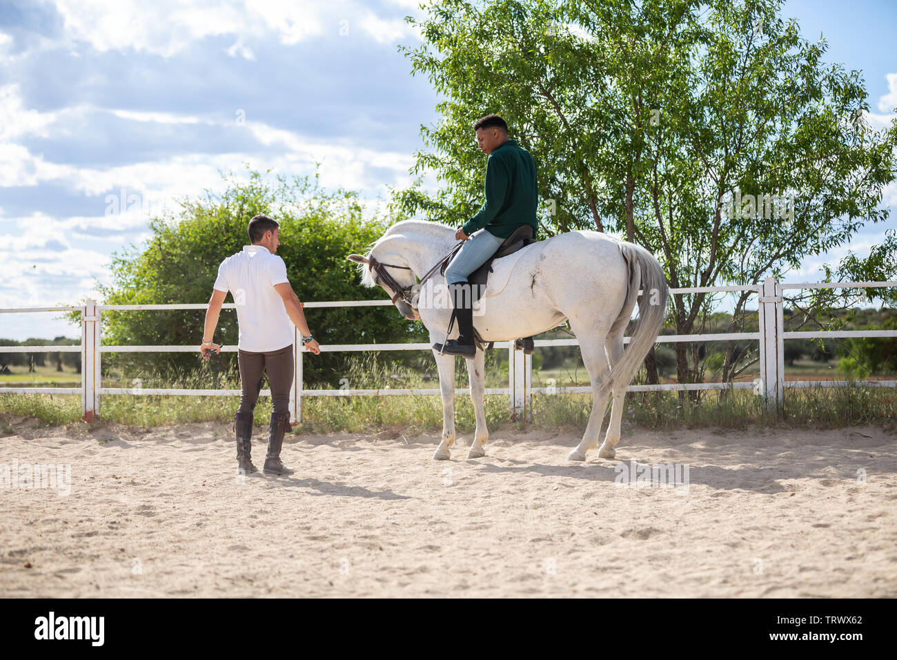 Young man teaching black guy to ride horse in paddock on cloudy day on ranch Stock Photo