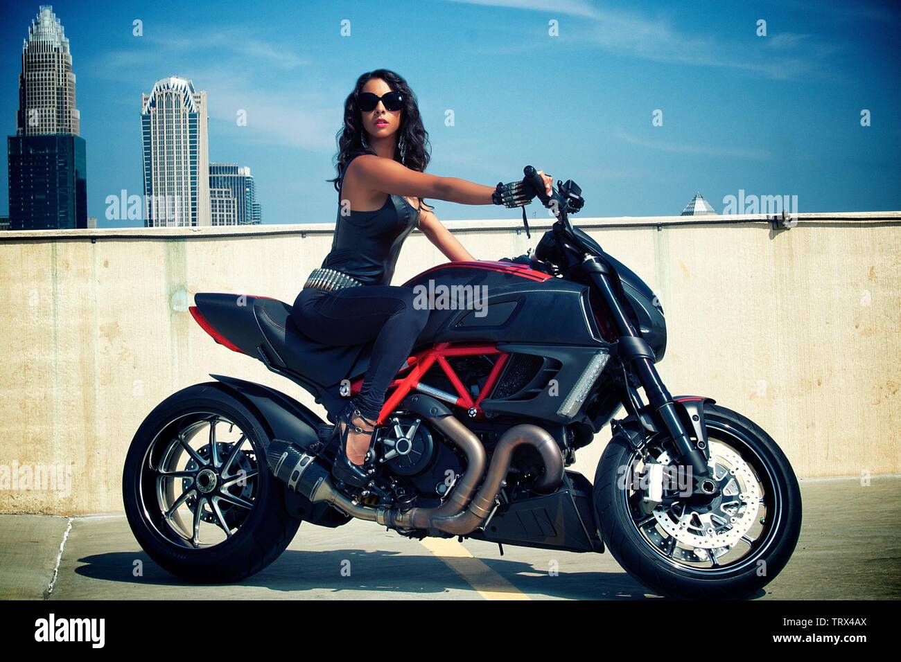 Black Woman on Ducati Motorbike with Skyline in the Background Wearing All Black and Sunglasses Shades Stock Photo