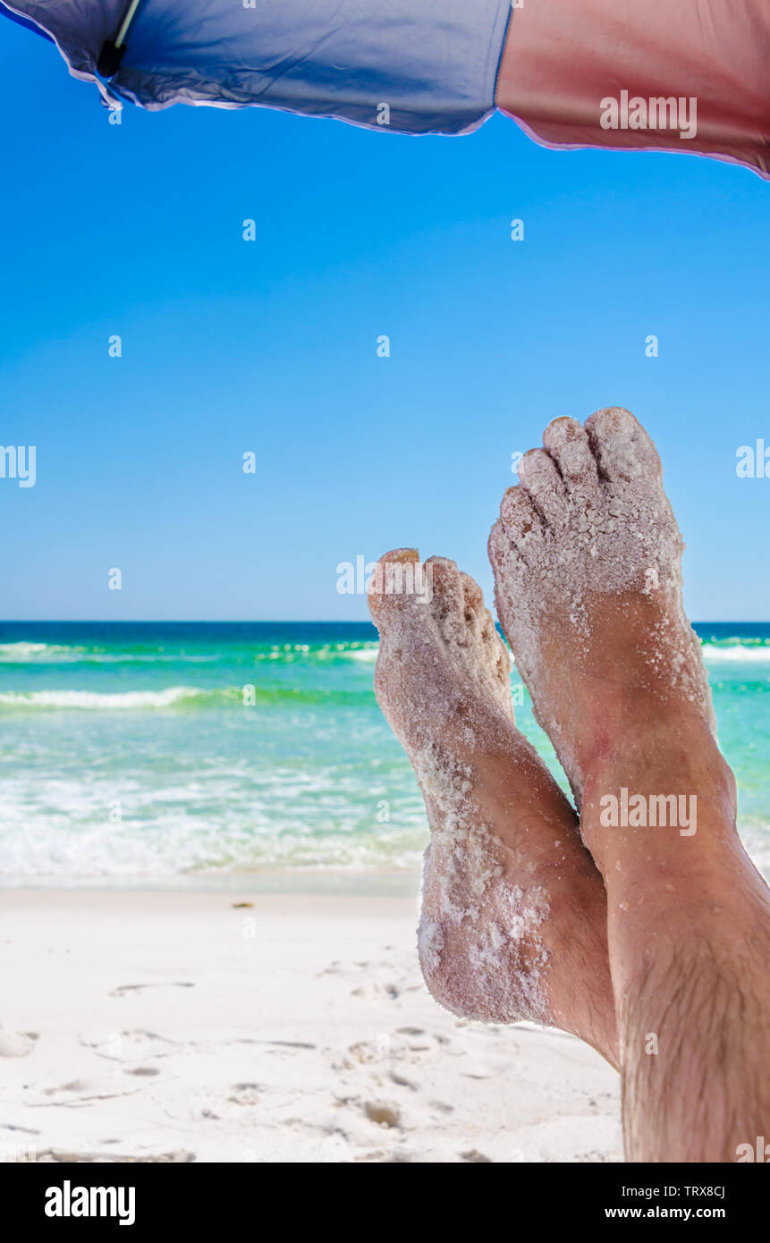 Man's sandy feet under umbrella, with ocean beach waters in the background. Beach image depicting relaxing and enjoying nature. Stock Photo