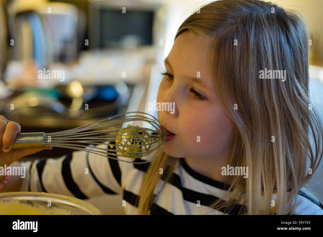 Young Girl Licking Cake Mix From A Metal Whisk In A Kitchen With Bokeh