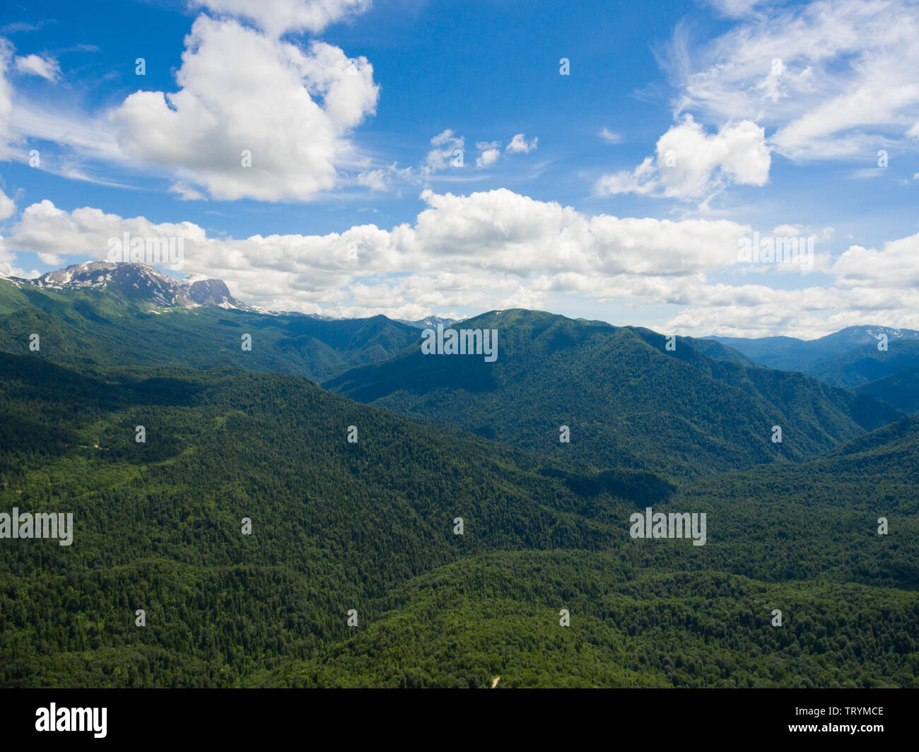Aerial photo. Summer landscape with mountain peaks covered with snow, cloudy sky and a segment of the road through the oak forest. Stock Photo