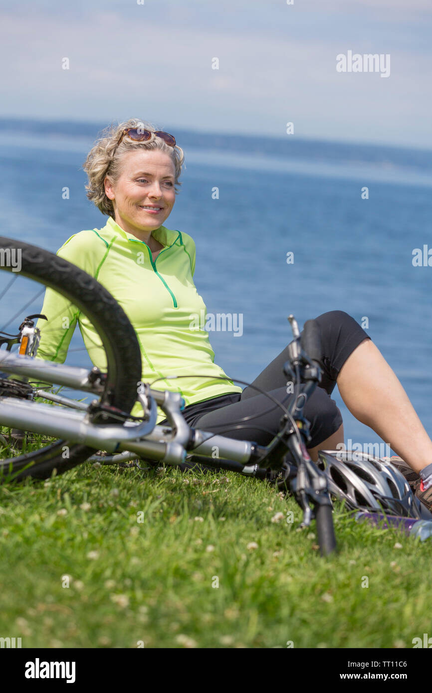 Beautiful, active, athletic, healthy middle-aged mature woman with bicycle sitting on grass outdoors. Stock Photo
