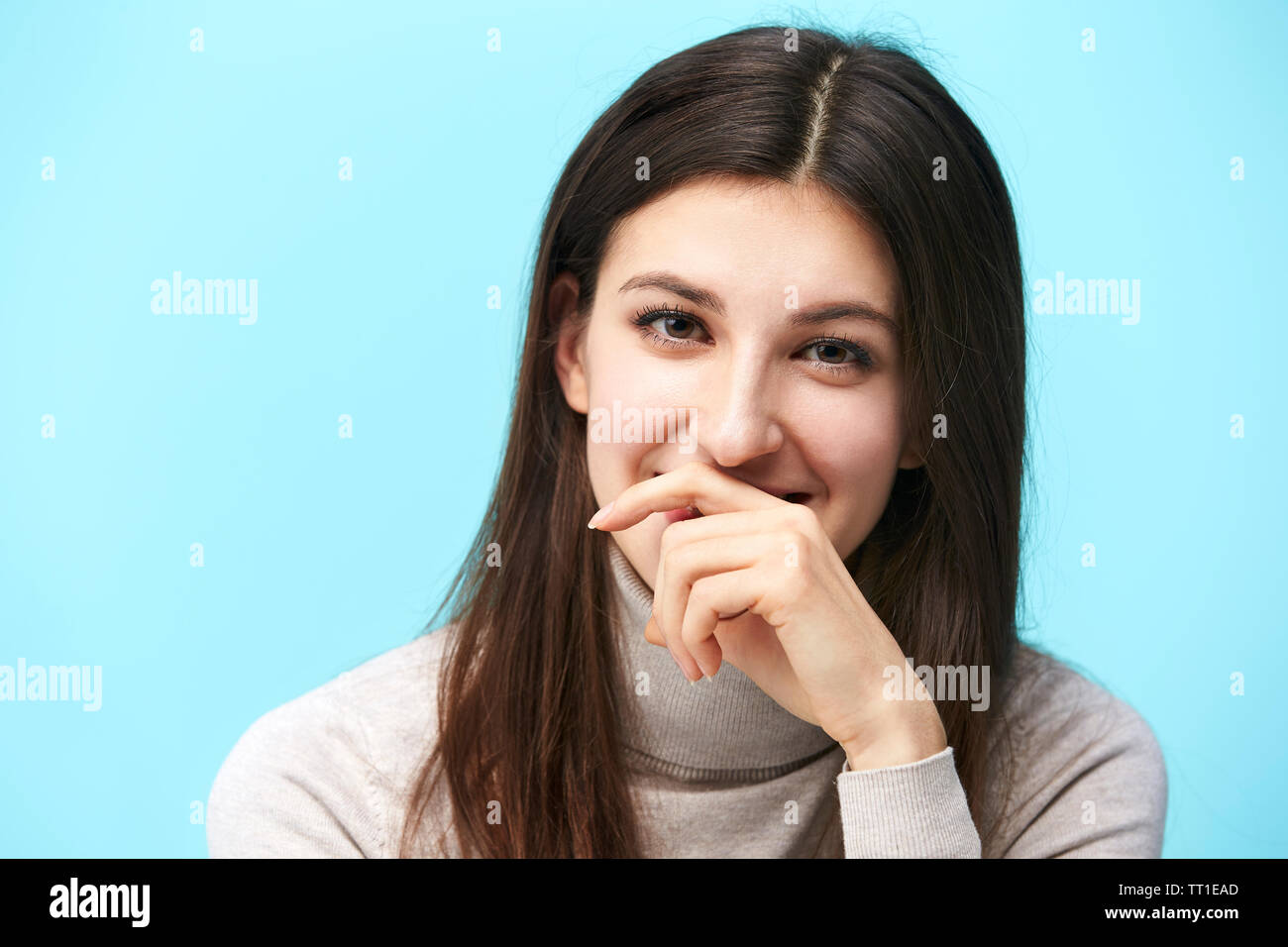 portrait of a beautiful young caucasian woman, head shot, looking at camera smiling, hand covering mouth, isolated on blue background Stock Photo