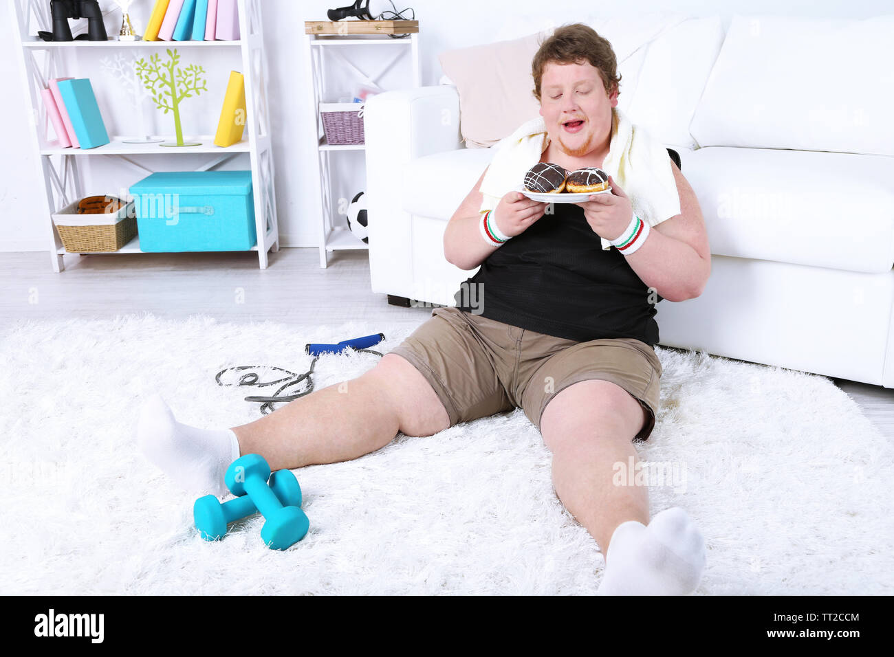 Large fitness man eating unhealthy food and trying to take exercise  at home Stock Photo