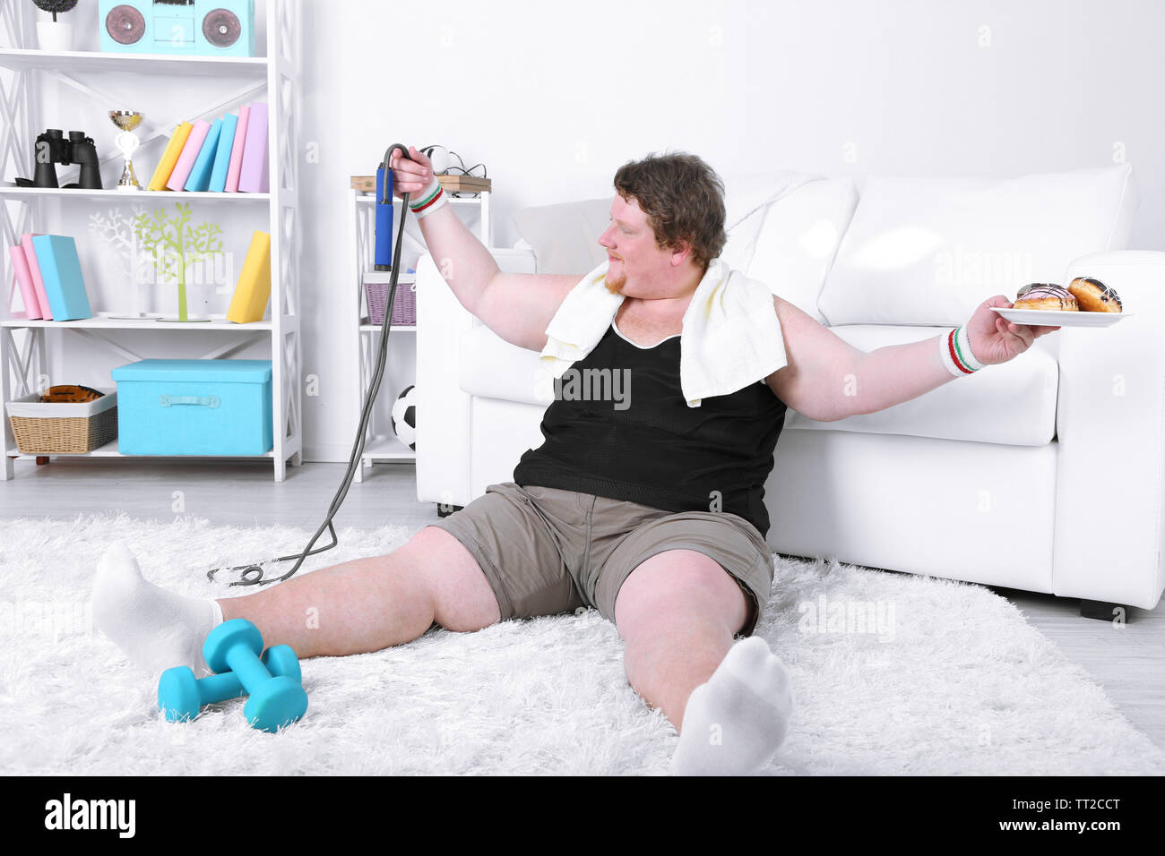 Large fitness man eating unhealthy food and trying to take exercise  at home Stock Photo