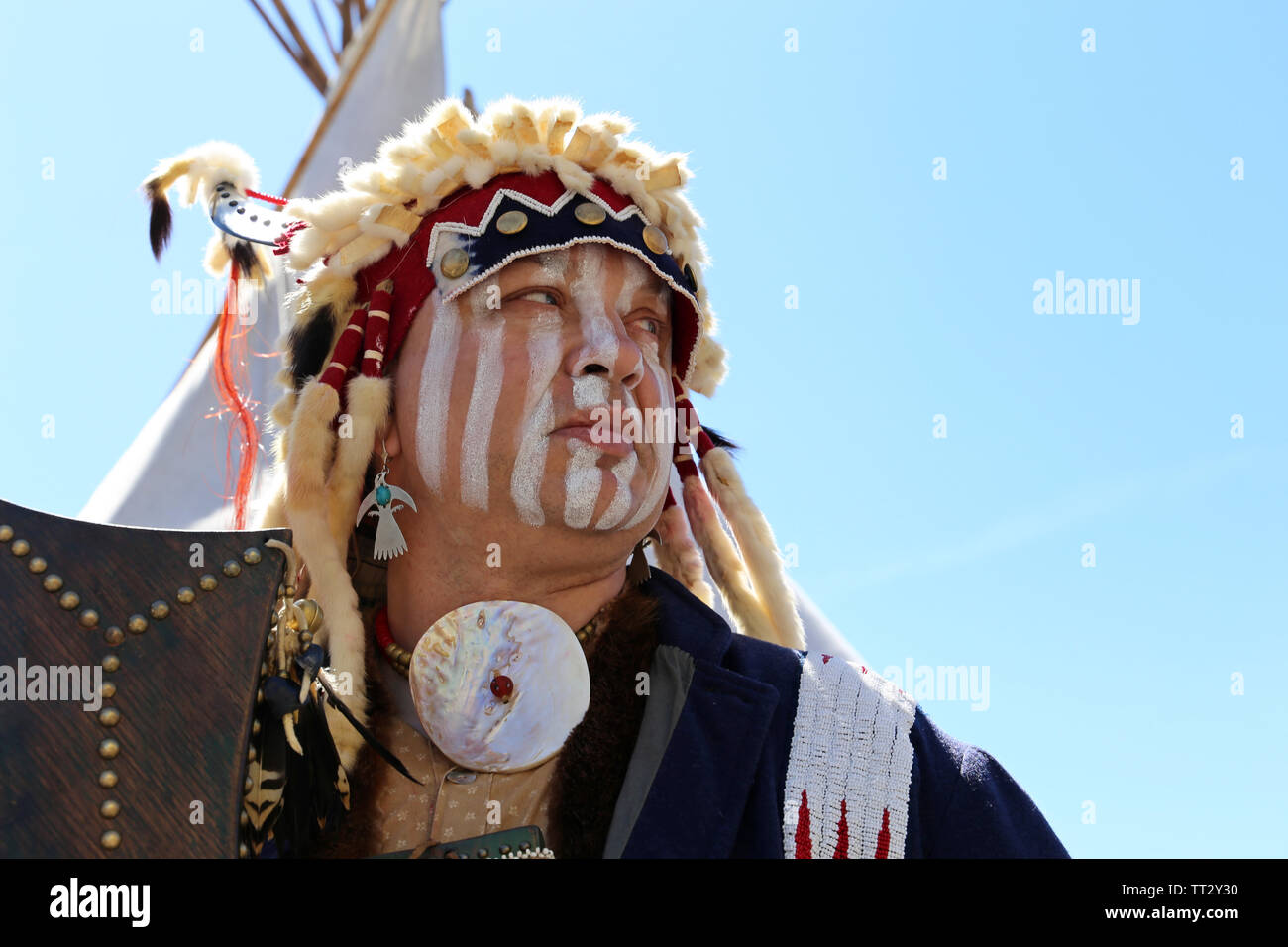 A man in the war paint and clothes of a North American Indian stands with weapon near a wigwam against the blue sky. Reconstruction of native american Stock Photo