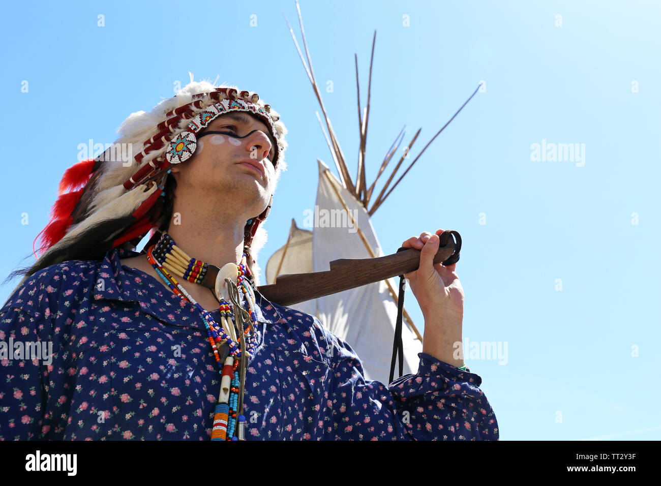 A man in the war paint and clothes of a North American Indian stands with weapon near a wigwam against the blue sky. Reconstruction of native american Stock Photo