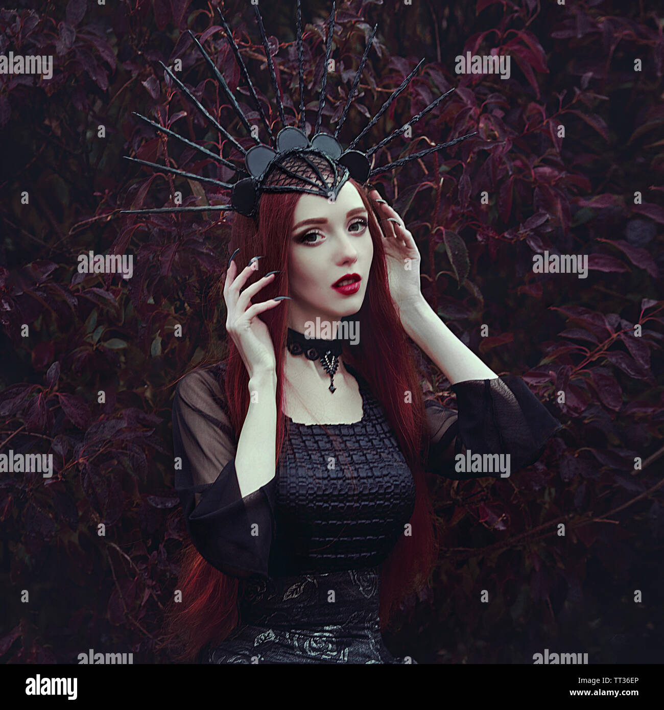 A beautiful woman with pale skin and long red hair in a black dress and in the black crownk. Girl witch with vampire claws and red lips. Gothic look. Stock Photo