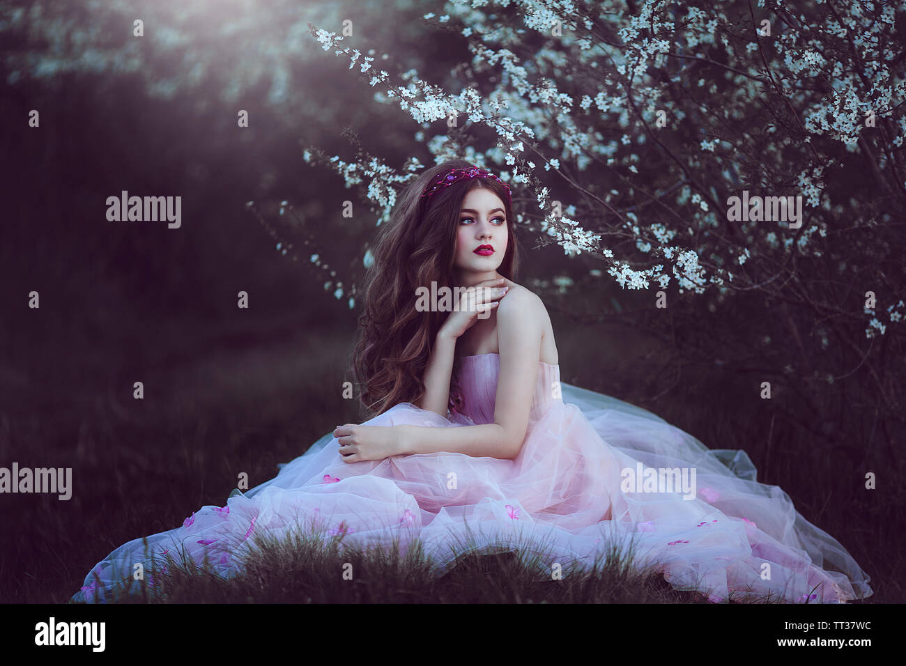 Beautiful Romantic Girl with long hair in fairy long pink dress sitting near flowering tree.Fantasy art. Creative colors and Artistic processing. Stock Photo