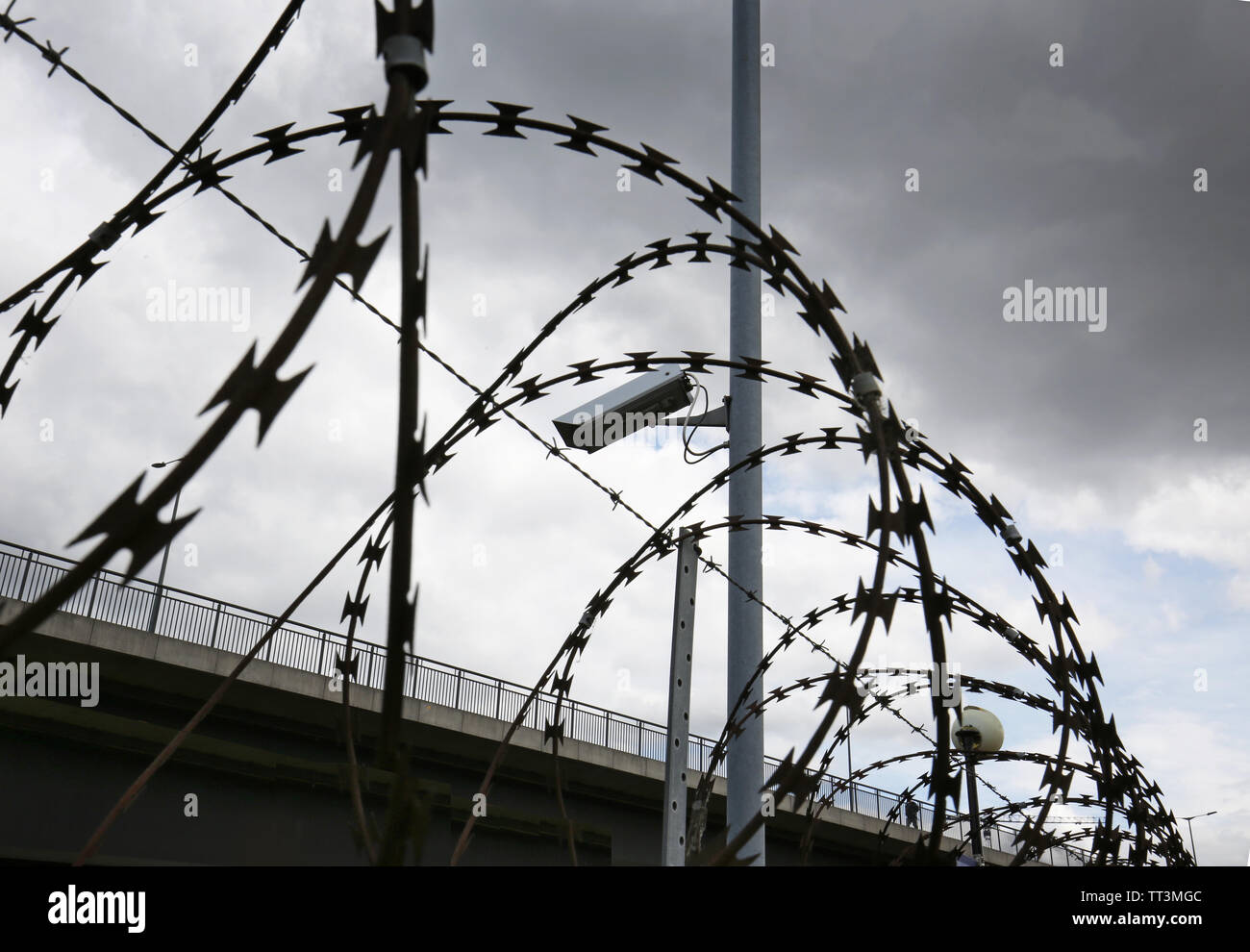 Close-up of razor wire and barbed wire surounding a secure compound in east London, UK. Shows security camera and lighting. Road viduct beyond. Stock Photo