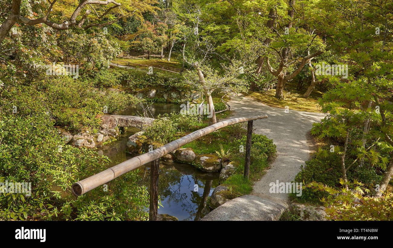 Japanese garden with a stone bridge across a river.  A path leads into the bushes. Stock Photo
