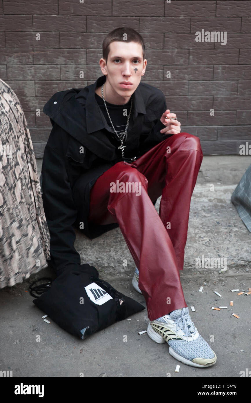 Moscow, RUSSIA - June 12, 2019: A young man in a black jacket, burgundy leather pants, and a cross-shaped tattoo on his face is sitting on the sidewal Stock Photo