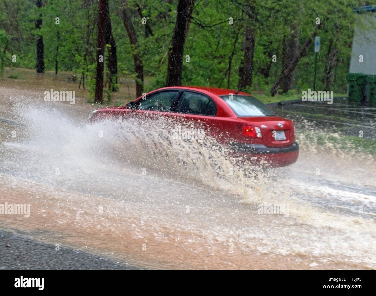 A red car splashes through dangerously high water driving on a back road as a river floods it