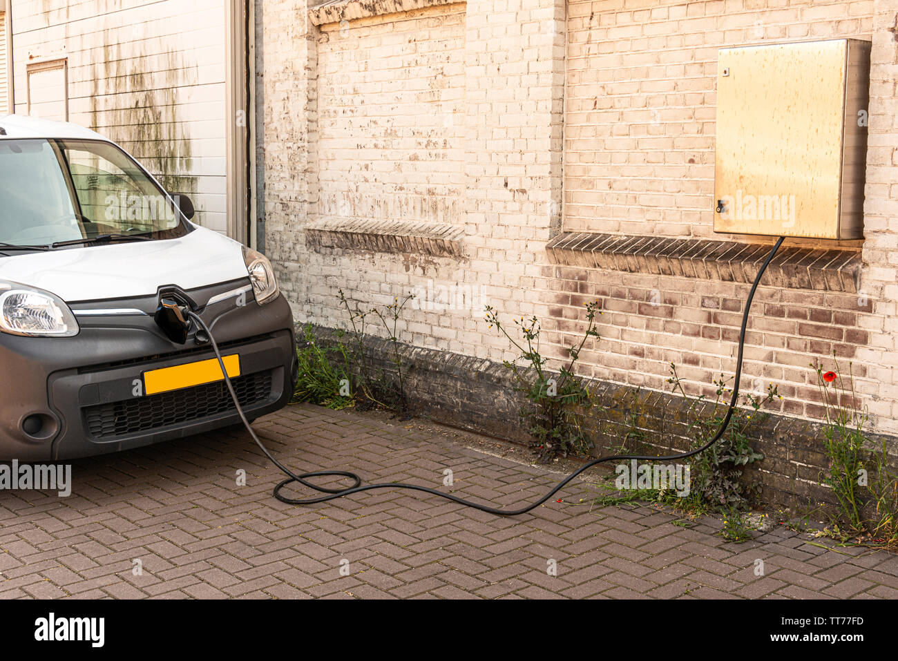 white black colored car standing next to a wall is electrically charged via a cable connected to a connection point hanging on a stone wall Stock Photo