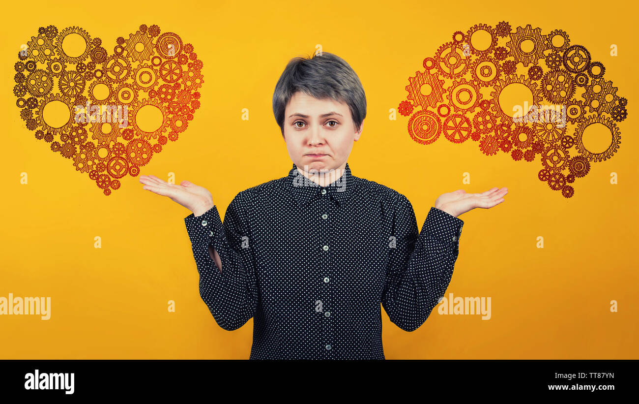 Perplexed woman balancing outstretched hands having hesitation shrugging shoulders as heart and brain symbol on different sides over yellow wall. Diff Stock Photo