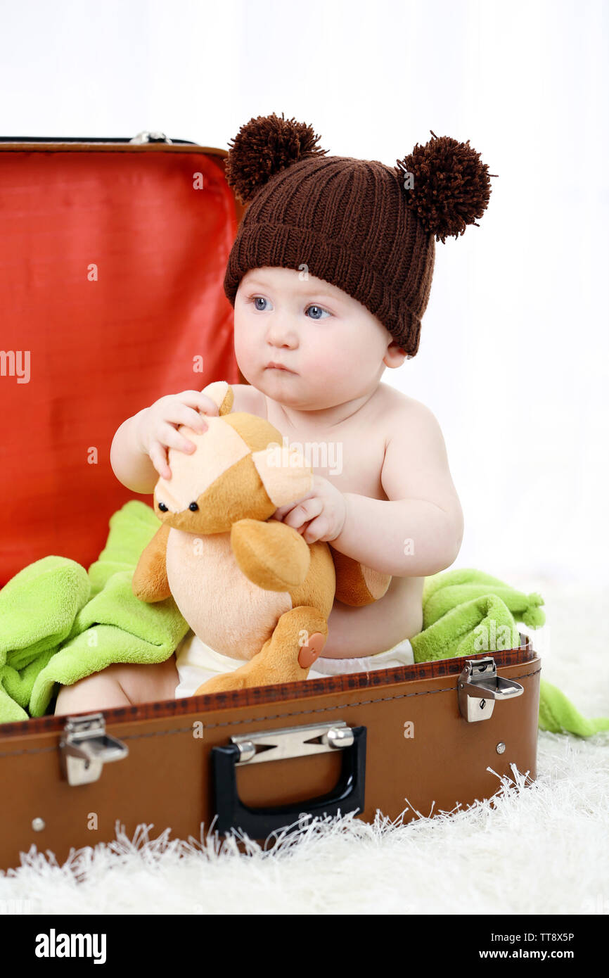 Cute baby boy in funny cap sitting in big suitcase on carpet, on light background Stock Photo