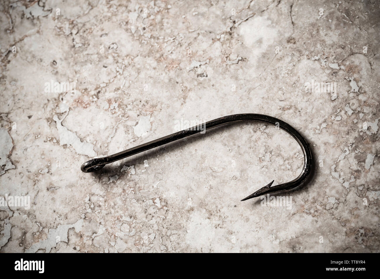 A single large seafishing hook displayed on a stone background. Hooks are equipped with barbs to stop them falling out of a hooked fish and to prevent Stock Photo