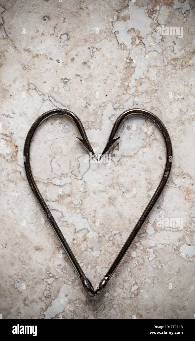 Two large, barbed seafishing hooks placed in the form of a heart and displayed on a stone background. Hooks are equipped with barbs to stop them falli Stock Photo