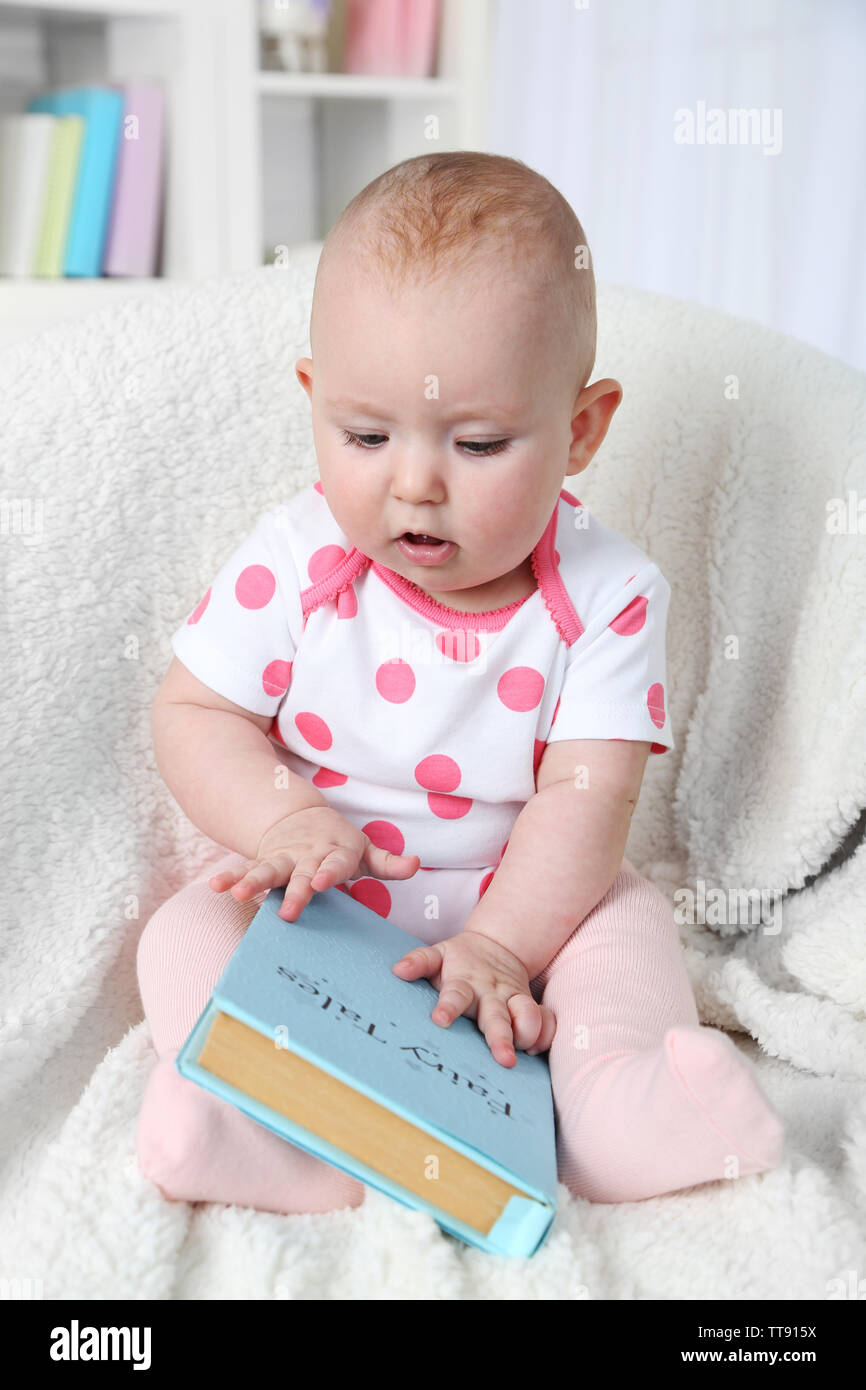 Cute baby girl sitting in armchair with book, on home interior