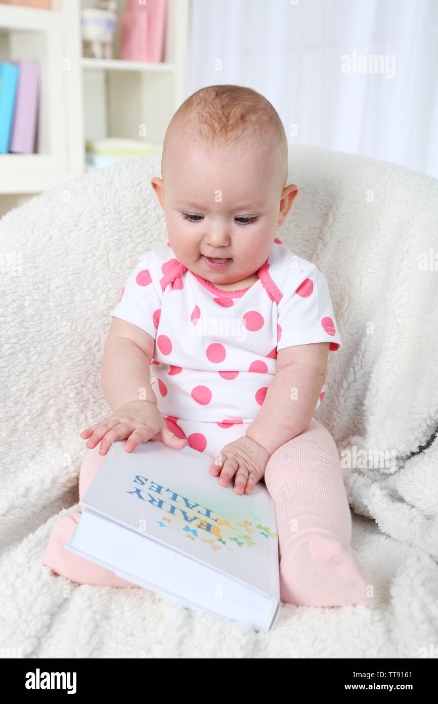 Cute baby girl sitting in armchair with book, on home interior