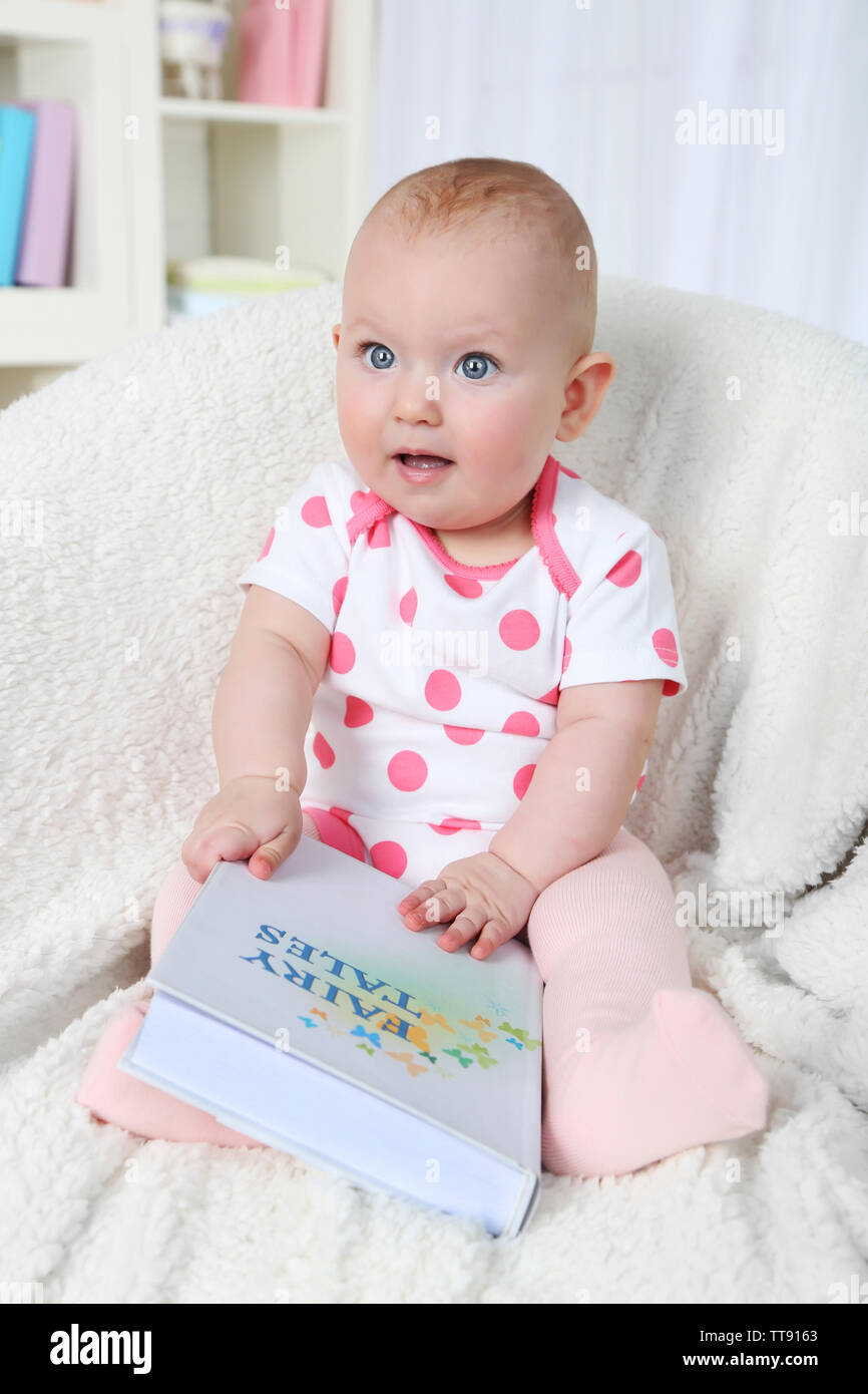 Cute baby girl sitting in armchair with book, on home interior
