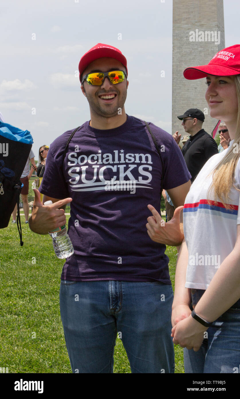 Washington, D.C., USA. 1st June, 2019. With the Washington Monument in the background at the National March To Impeach on 1 June, 2019, a grinning man faces the camera, wearing a red 'Make America Great Again' baseball cap/hat, and points with both hands, one holding a bottle of water, to the words 'Socialism Sucks' printed on his t-shirt. The woman beside him, partially visible, wears the same cap and smiles slightly while facing left. They were two of several pro-Trump counterprotesters at the People Demand Action-organized demonstration on the Washington Monument Grounds. Kay Howell/Alamy Stock Photo